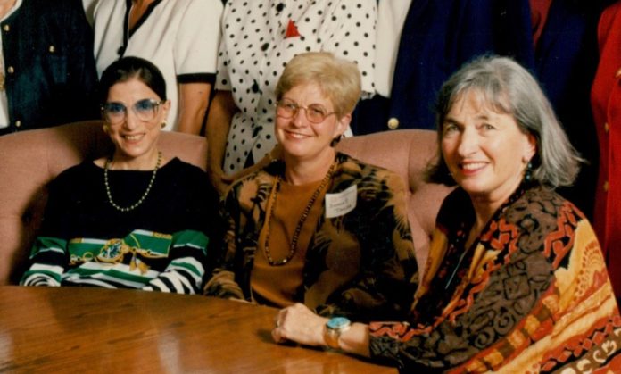 Three middle aged white women sit in chairs around a roundtable smiling.