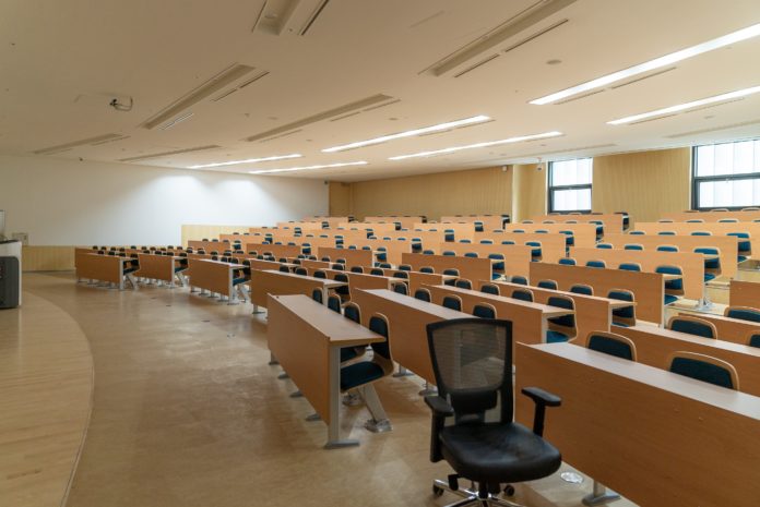 A large classroom is filled with rows of tables with chairs in front of them. After each row, the brown tables and chairs become more elevated.