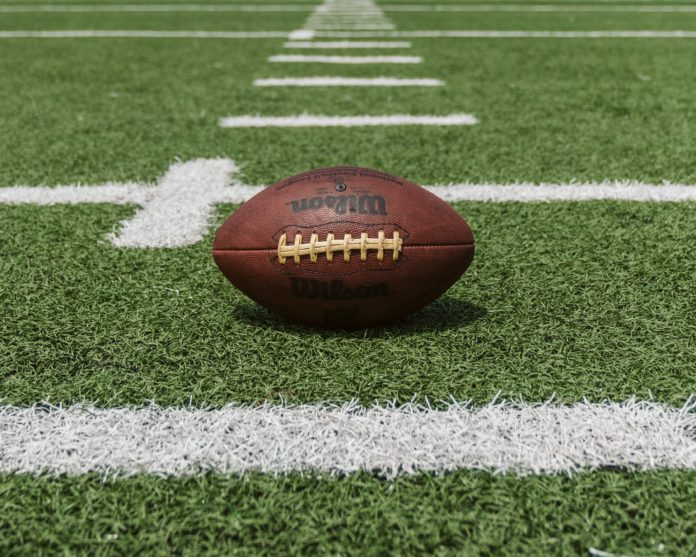 A brown football rests between yard lines on a football field. The brand name Wilson can be read on the football.
