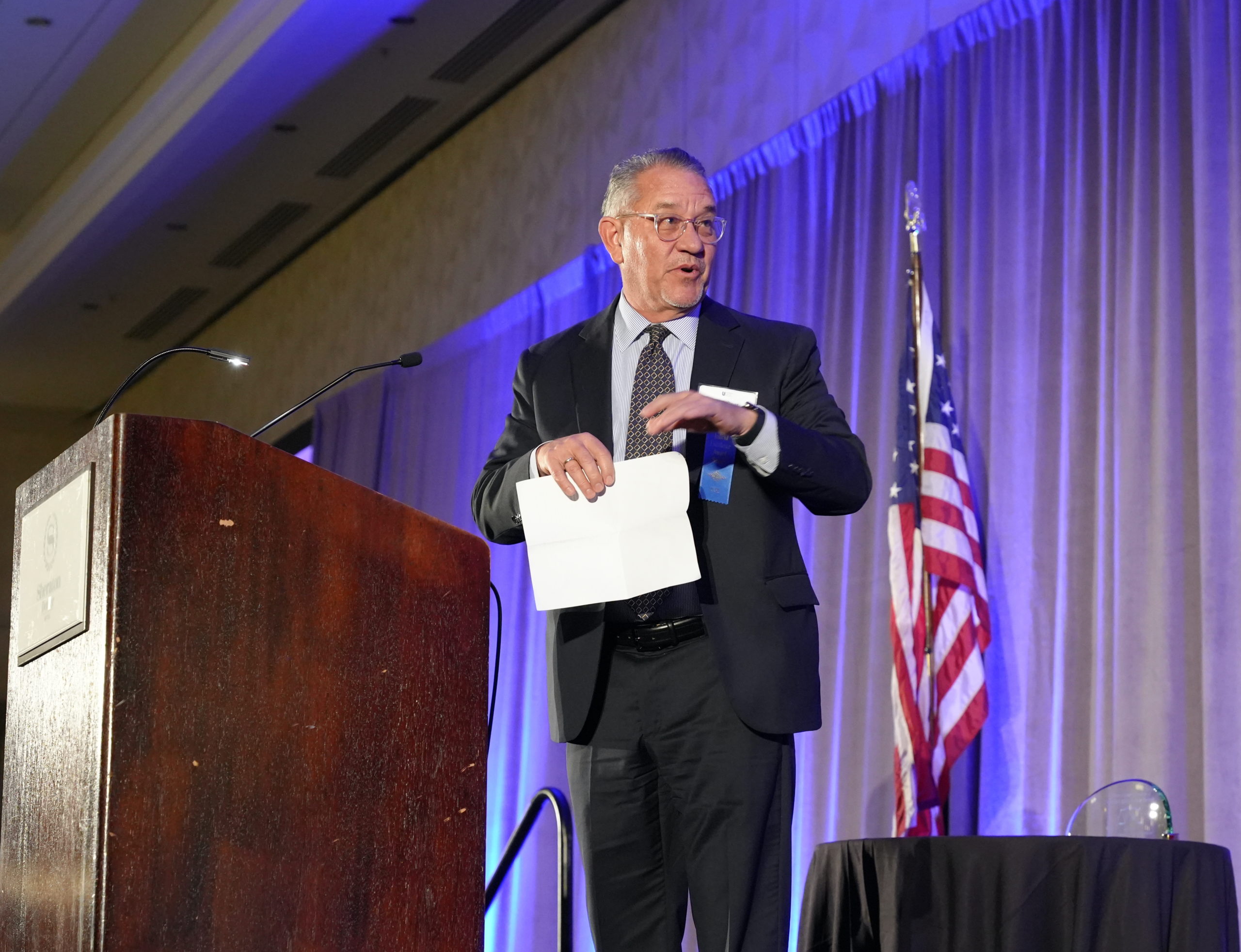 A man with gray hair and clear glasses in a black suit holds a piece of paper on a stage near a red podium with an American flag behind him and a table with a glass award and looks at someone off stage. 