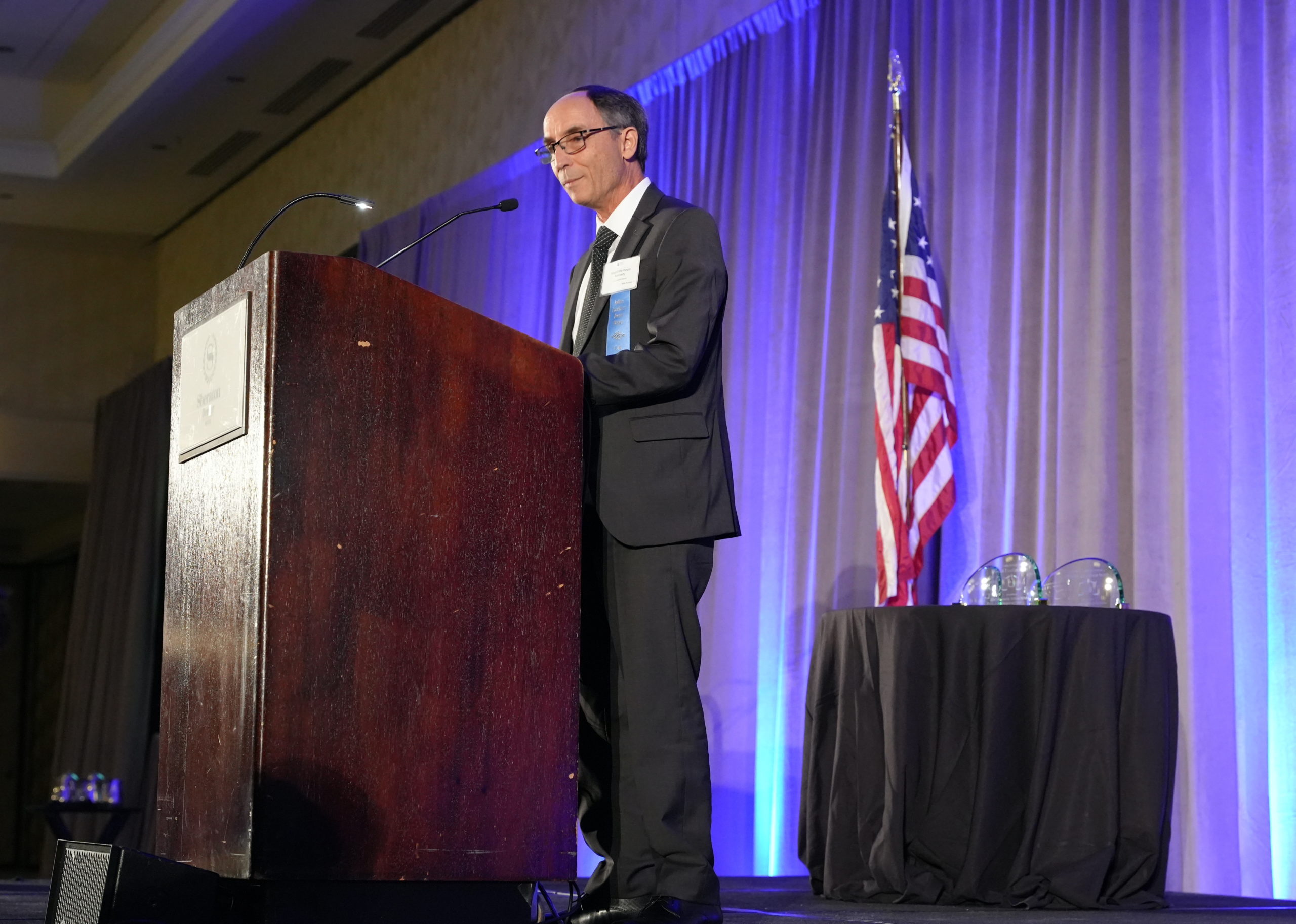 A balding man with dark hair wears a gray suit and stands in front of a red podium on a stage with the American flag behind him and a table with two glass awards.