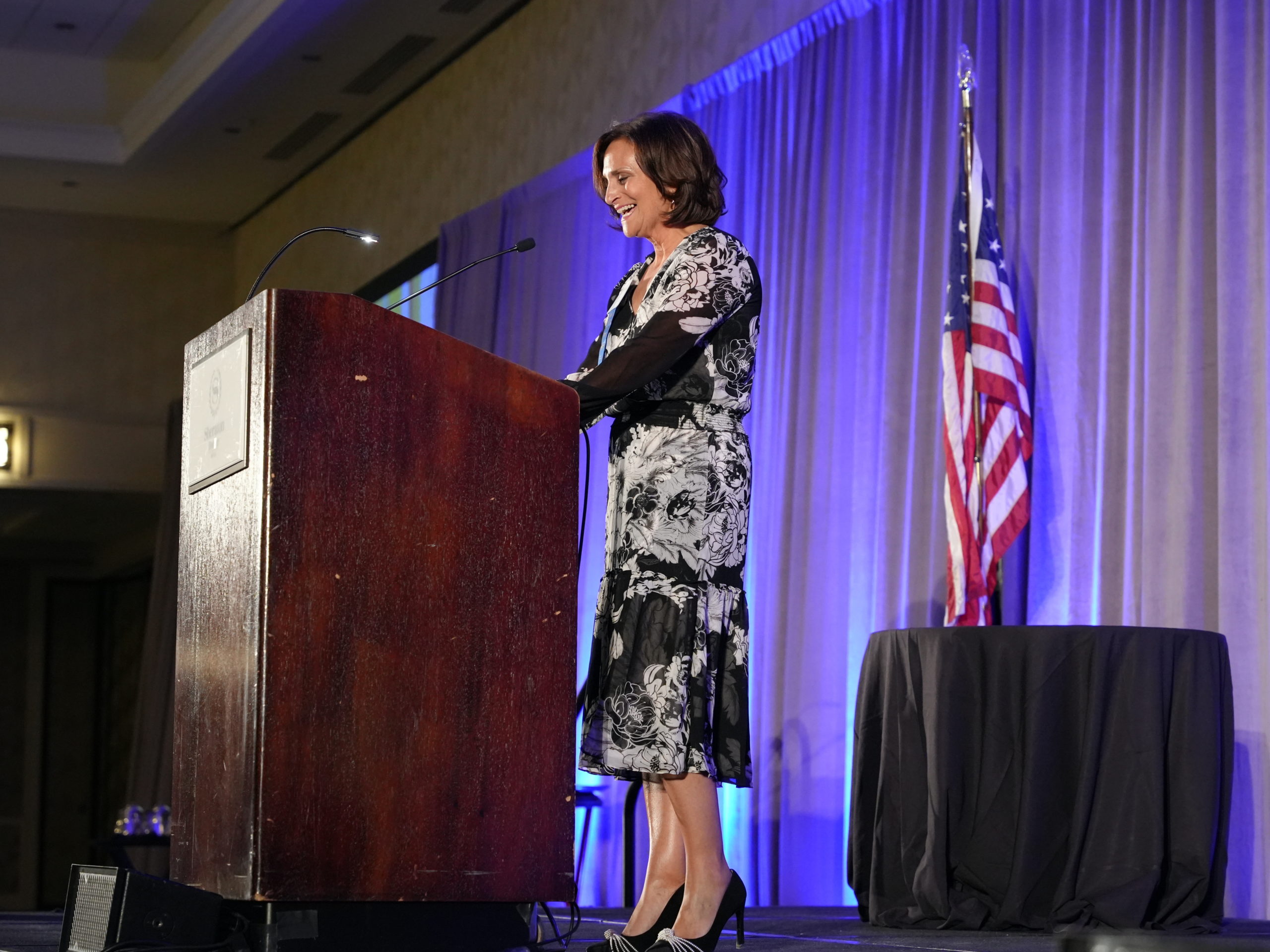 A woman with short brown hair wears a patterned black and white dress and high heels and smiles and speaks in front of a red podium near an American flag.