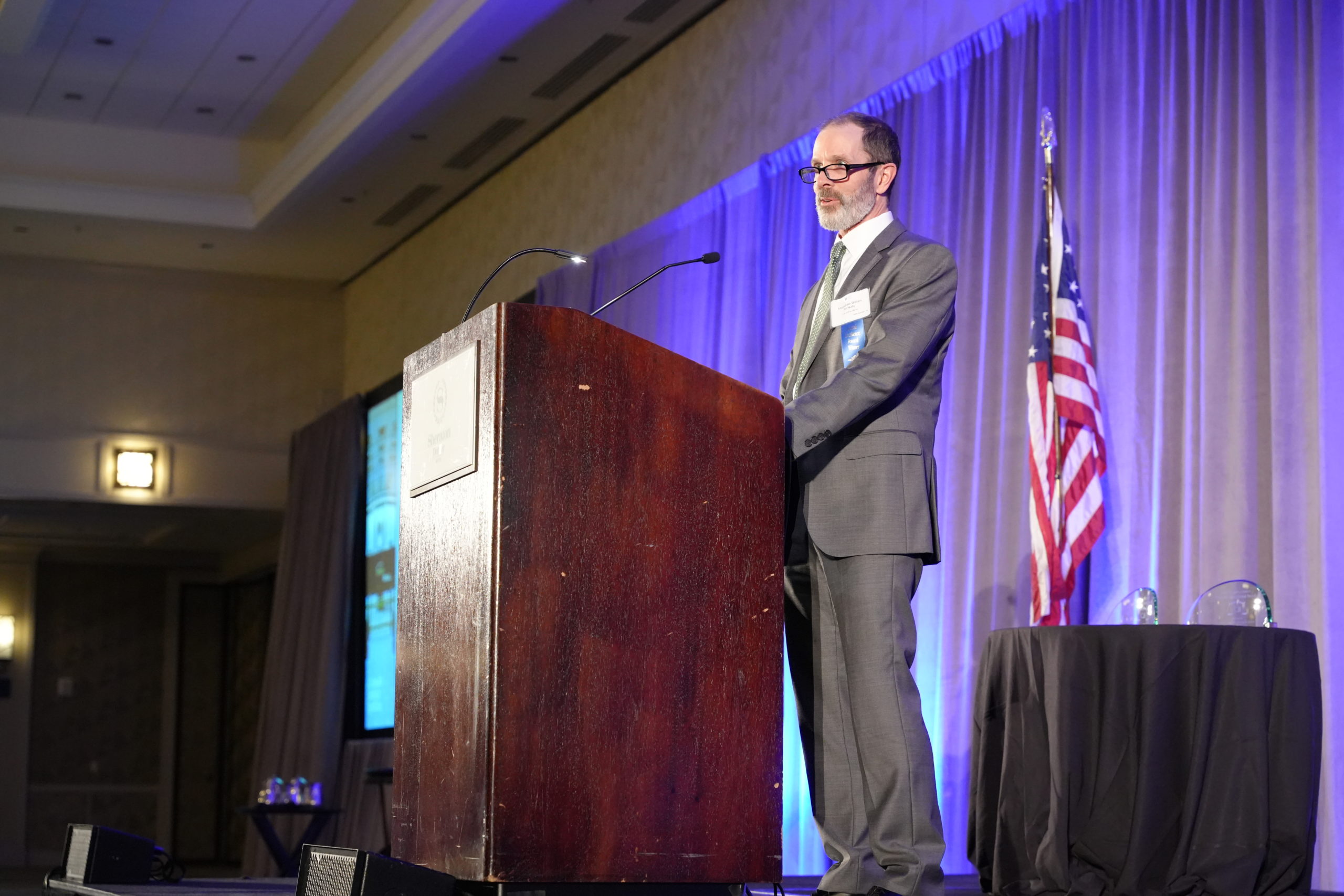  A man in a gray suit with dark hair and a gray beard wearing glasses stands in front of a red podium on a stage with the American flag behind him and a table with two glass awards. 