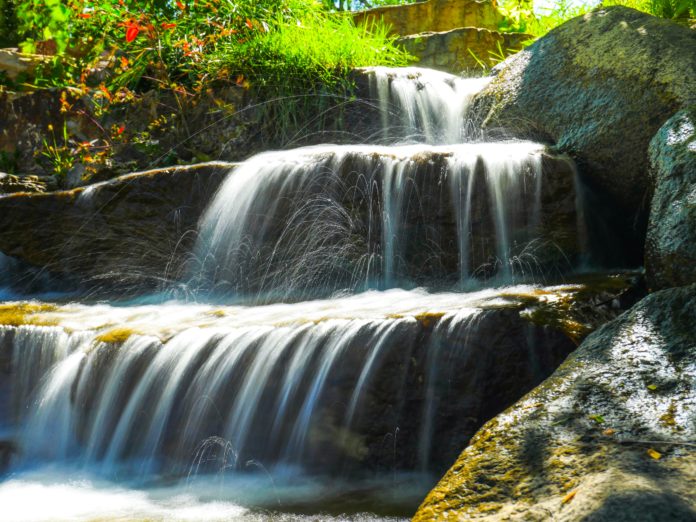 pablo-E2JB1m-8Ix8-unsplash A tributary feeds into the Arkansas River near a bridge. Water is falling over three different rock levels with lush green plants peaking over the bank on one side.