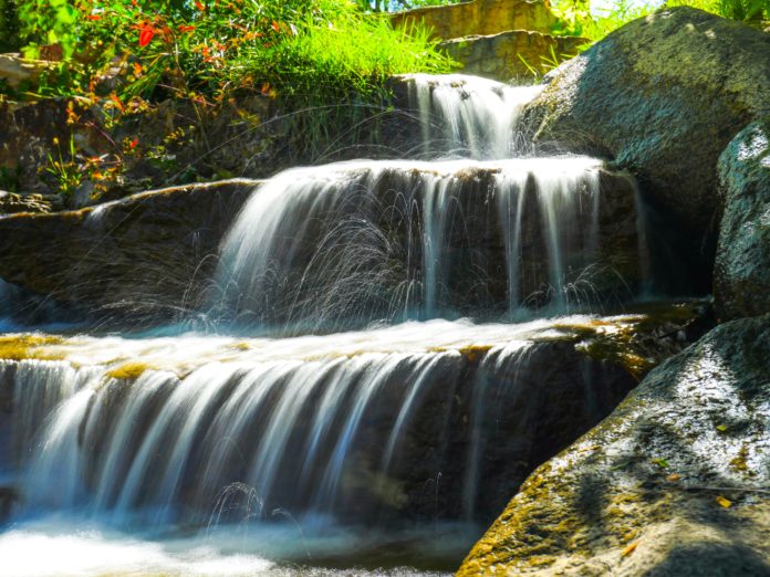 A tributary feeds into the Arkansas River near a bridge. Water is falling over three different rock levels with lush green plants peaking over the bank on one side.