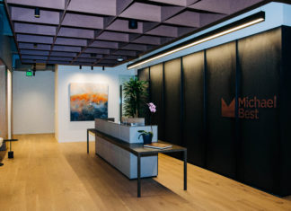 Michael Best’s New Denver Space is Set for a Growing Team A wide shot of a modern office lobby with tall ceilings and hardwood floors. A back wall includes the logo and name Michael Best. In the center is a modern reception table with a computer and orchid.