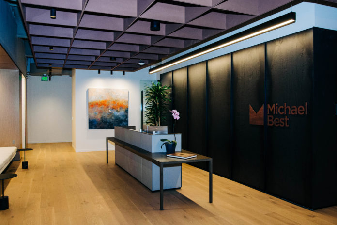 A wide shot of a modern office lobby with tall ceilings and hardwood floors. A back wall includes the logo and name Michael Best. In the center is a modern reception table with a computer and orchid.