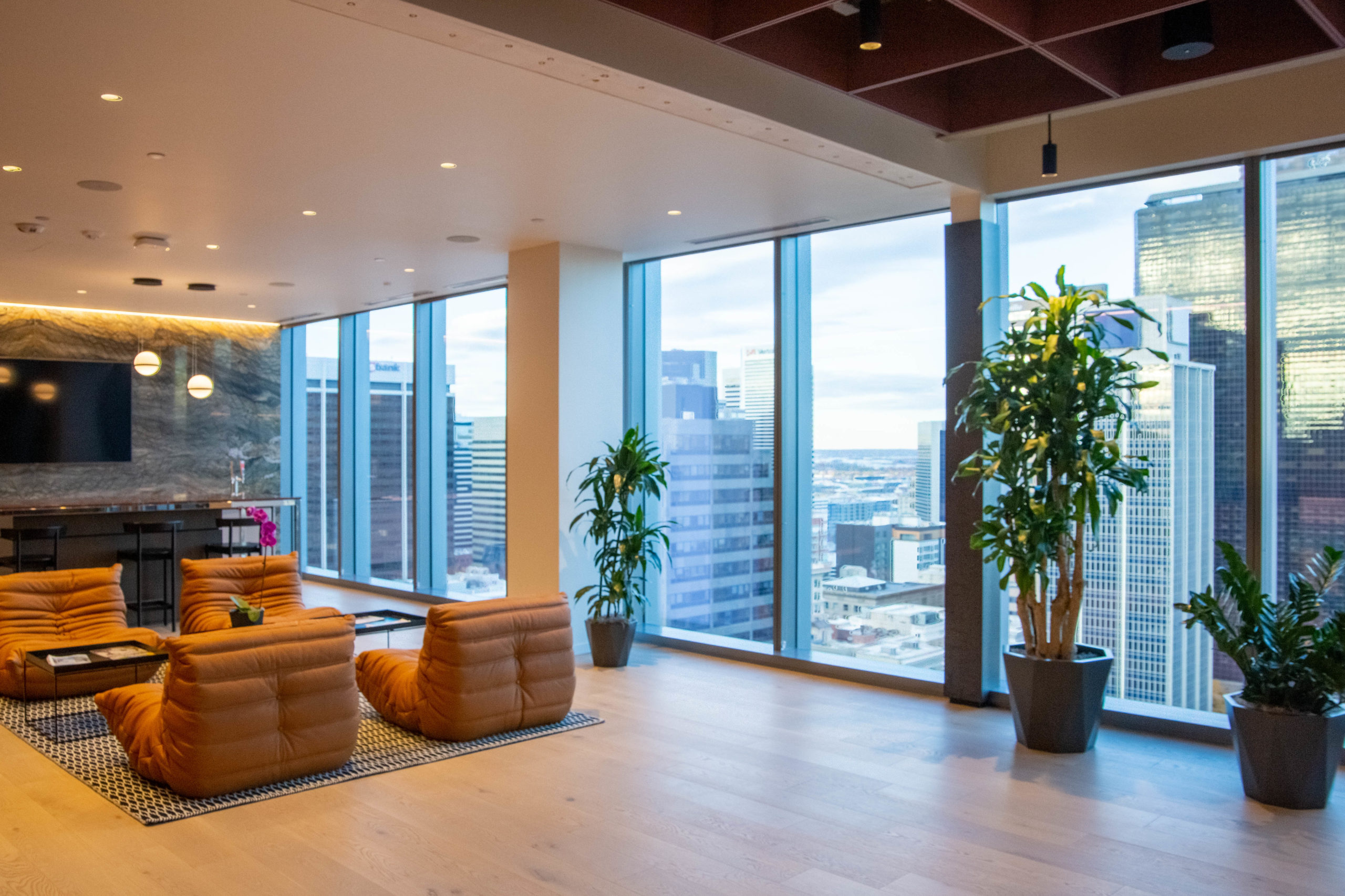 A large open room with floor to ceiling glass windows overlooking a city. Inside the room, four modern orange chairs face each other. In the background, bar stools sit around a counter and marble wall with a television.