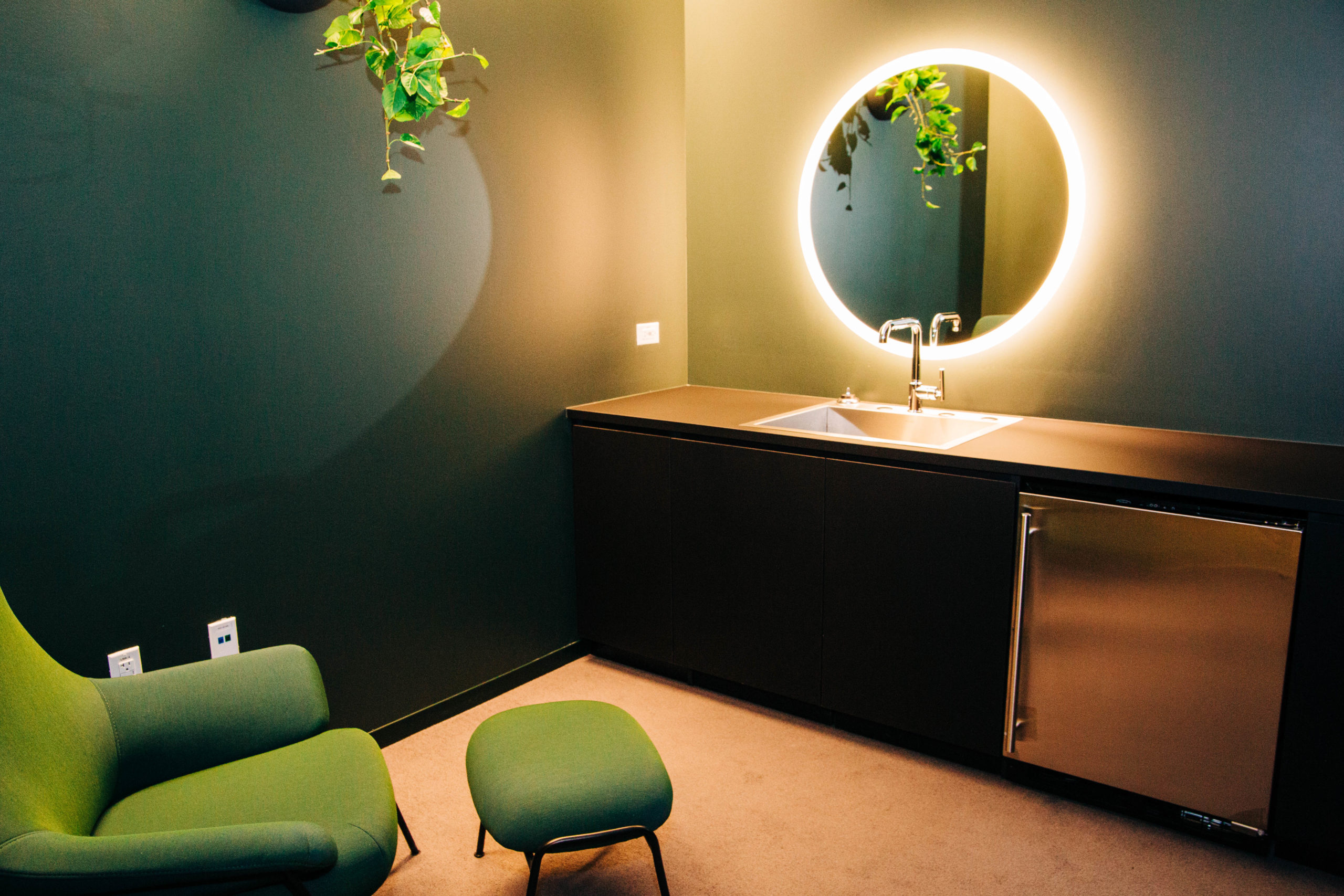 A small room painted dark green with soft lighting. One side includes a counter with a metal door, a sink and mirror with lights around it. Nearby is a modern green arm chair and ottoman. 