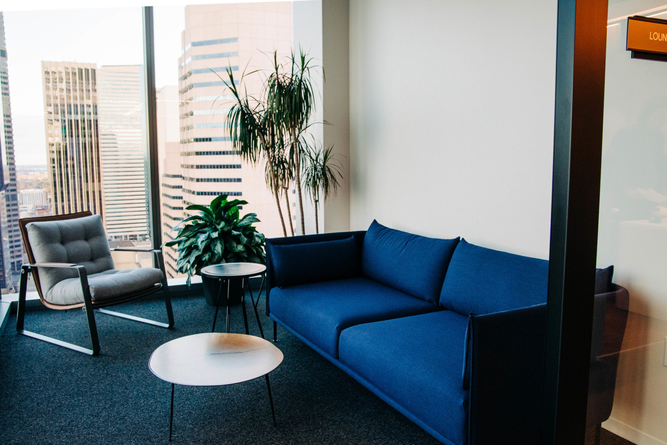 A modern blue couch and modern gray chair face each other with a coffee table in the middle of a room with floor to ceiling glass on two sides overlooking a city. 