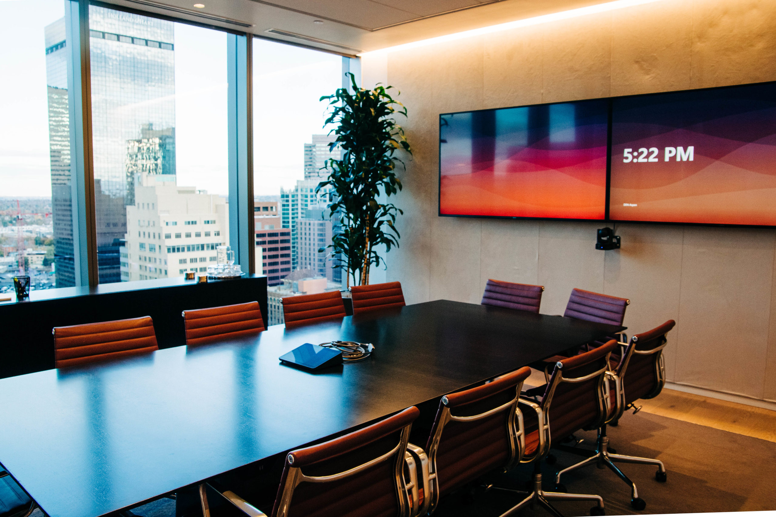 A large black table with red office chairs around it in a modern conference room with floor to ceiling windows overlooking the city. 