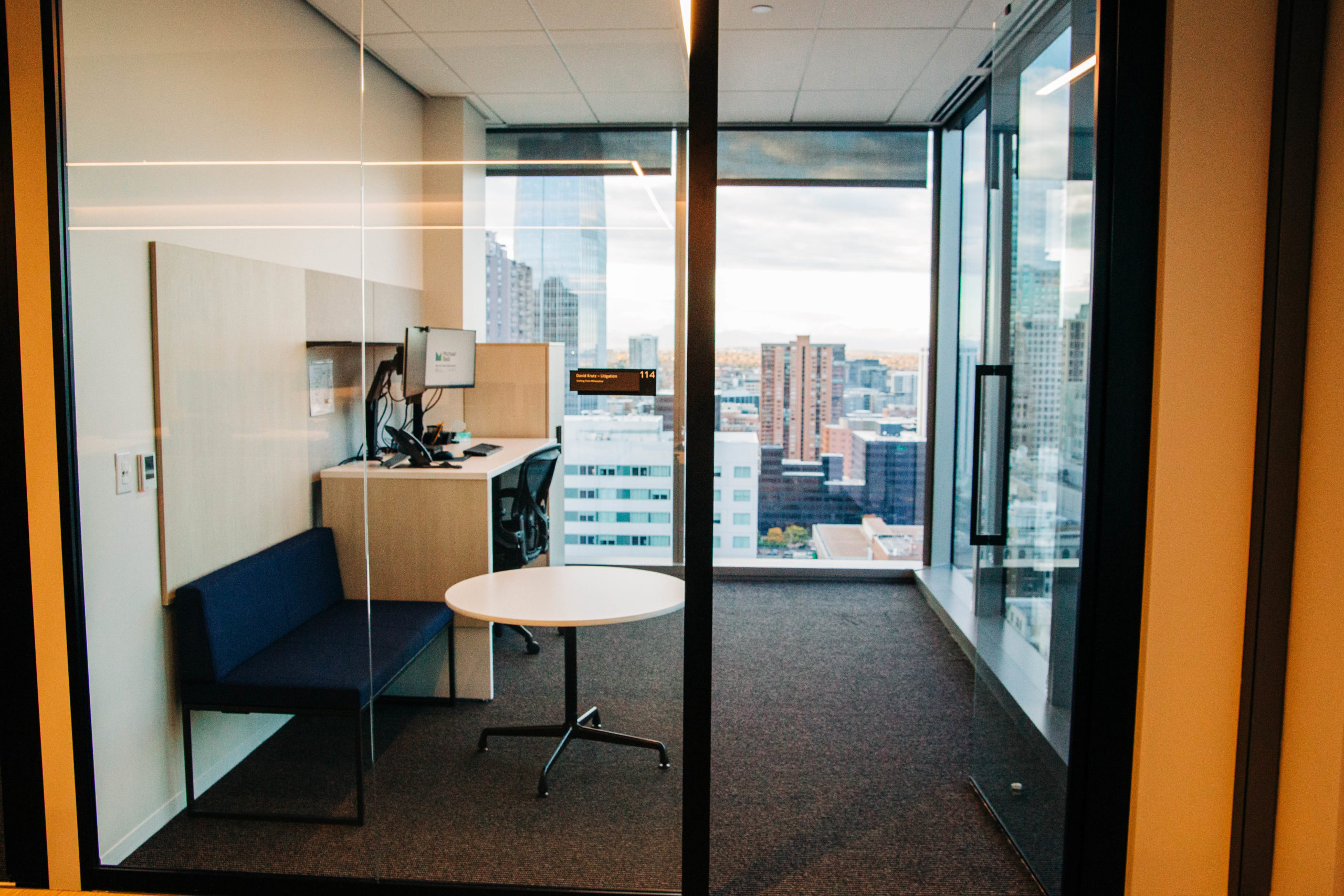 An empty modern office with floor to ceiling windows, a standing desk, small couch and gray carpet. 