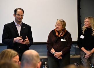 Colorado AG Announces Natural Resources Fellowship in Honor of Late Justice Hobbs A man with eye glasses wearing a suit coat stands next to two women in front of an audience of people. One of the women is wearing a brown top with eye glasses on top of her head while the other woman is wearing a black top. Behind them is a white backdrop.