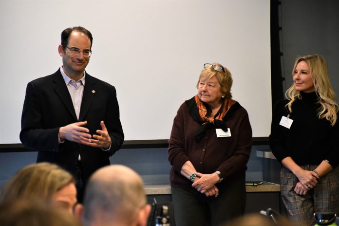 A man with eye glasses wearing a suit coat stands next to two women in front of an audience of people. One of the women is wearing a brown top with eye glasses on top of her head while the other woman is wearing a black top. Behind them is a white backdrop.