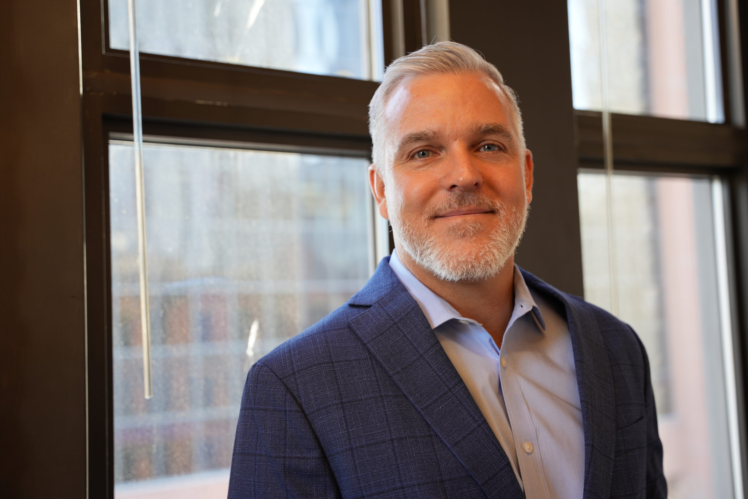 A man with gray hair, a gray beard and blue eyes in a blue suit smiles slightly in front of large windows. 