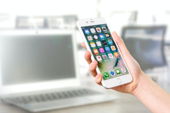 A hand holds a white iPhone with a home screen in front of a blurred background of an office and laptop.
