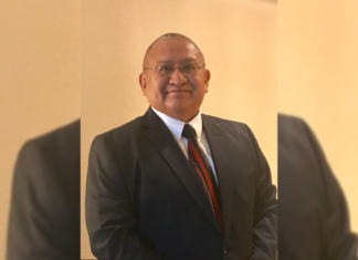 Director Begins Work in New Office Helping Investigate Missing, Murdered Indigenous Relatives A man stands with a suit on and red tie. He wears glasses and there’s a yellow background