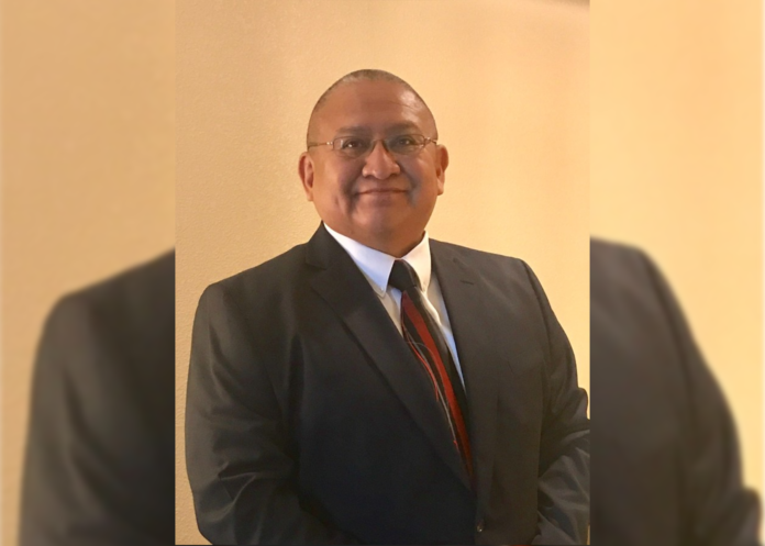 A man stands with a suit on and red tie. He wears glasses and there’s a yellow background