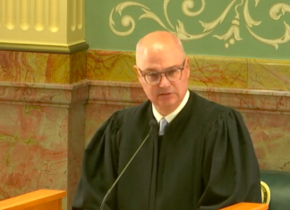 Boatright’s State of the Judiciary Outlines Workplace Changes in Colorado Courts An older bald man with round frame glasses wearing black judge’s robe speaks at a wood podium in front of an ornate wall with pink marble and green walls with decorative painting.