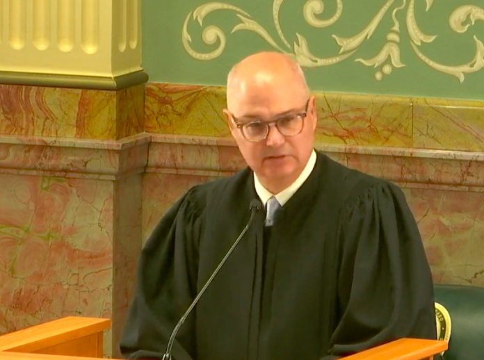 An older bald man with round frame glasses wearing black judge’s robe speaks at a wood podium in front of an ornate wall with pink marble and green walls with decorative painting.