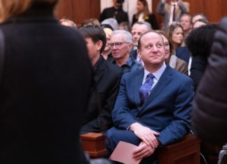 Gov. Polis Addresses Club Q Shooting, Housing in State of the State A man wearing a blue suit and tie sits in front of a large gathering of people.