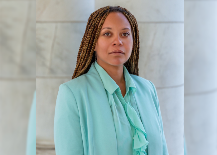 A Black woman with braids wearing a teal suit looks straight ahead in front of white pillars.