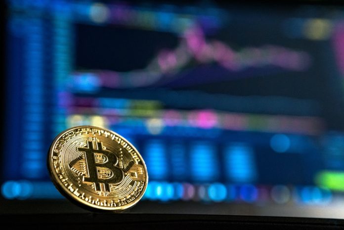 andre-francois-mckenzie-iGYiBhdNTpE-unsplash (1) A gold coin with the letter “B” with two lines going through it can be seen. In the background is a blurred out line chart graph on a computer screen.