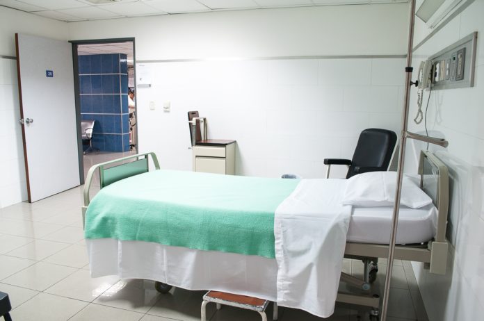 a hospital bed lies empty; just outside the room, health care workers are looking at files.