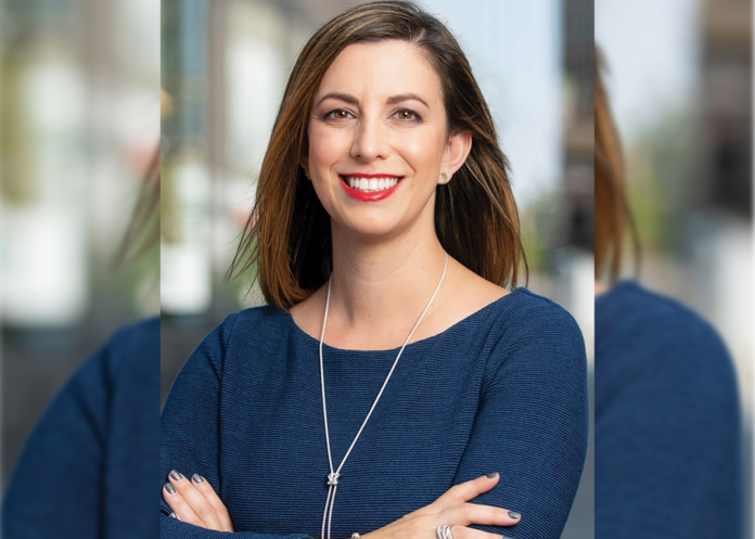 A woman wearing a blue top and necklace stands smiling with what appears to be a building behind her.