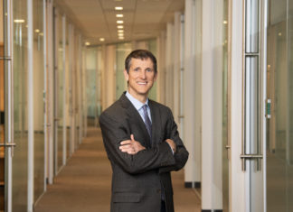 ACLU Colorado Names Arnold & Porter’s Tim Macdonald as Legal Director man with short hair and blue eyes wearing a gray suit smiles with his arms crossed in front of a long hallway in an office.