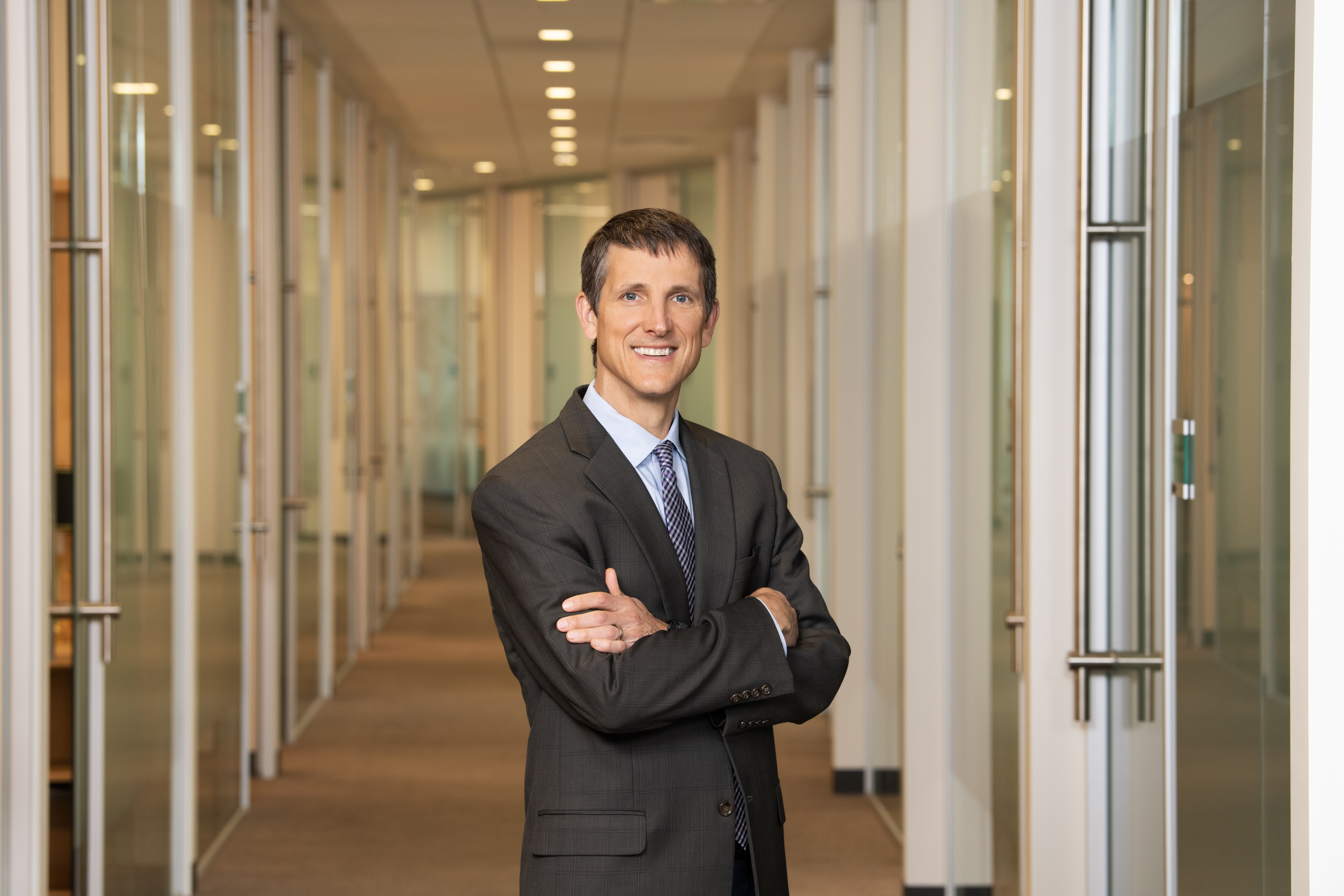 man with short hair and blue eyes wearing a gray suit smiles with his arms crossed in front of a long hallway in an office.