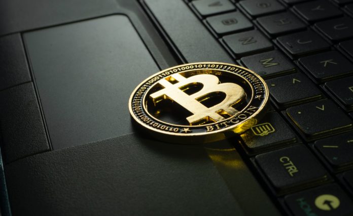 A gold coin has a “B” in the middle of it with two lines going through it which rests on a laptop keyboard. It reads “Bitcoin” on the bottom of the coin.