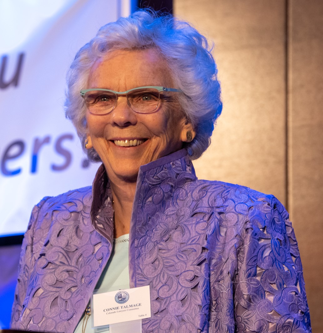 A woman with short white hair and blue frame glasses in a patterned purple suit top smiles beyond the camera. Behind her is a projector screen. She wears a name tag that reads Connie Talmage.