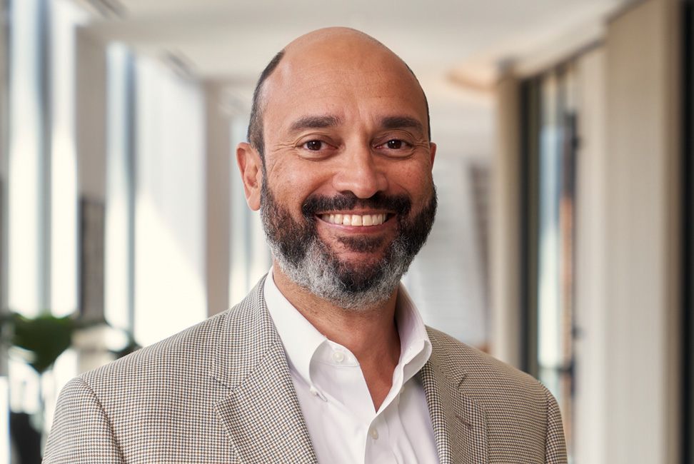 A middle aged Black man with brown eyes, a graying beard in a tan suit smiles in front of an office background. 