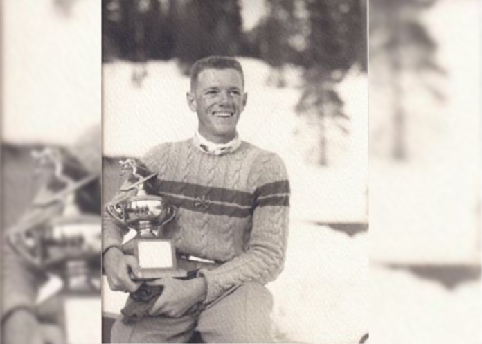 Gardner Paul Smith is smiling and holding a ski trophy and gloves while wearing a striped sweater with a collared white shirt underneath. The photo looks very old.