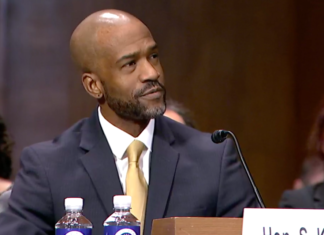 Senate Holds Vote and Hearing for Colorado Federal Court Nominees A middle aged bald Black man with a slight beard wearing a gray suit and yellow tie sits in front of a microphone at a table. He looks off to the right, appearing to listen to someone off camera.