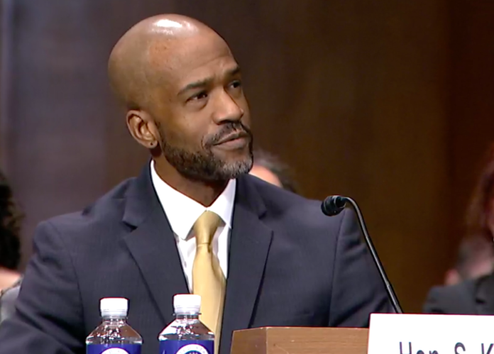 Kato Crews confirmation A middle aged bald Black man with a slight beard wearing a gray suit and yellow tie sits in front of a microphone at a table. He looks off to the right, appearing to listen to someone off camera.