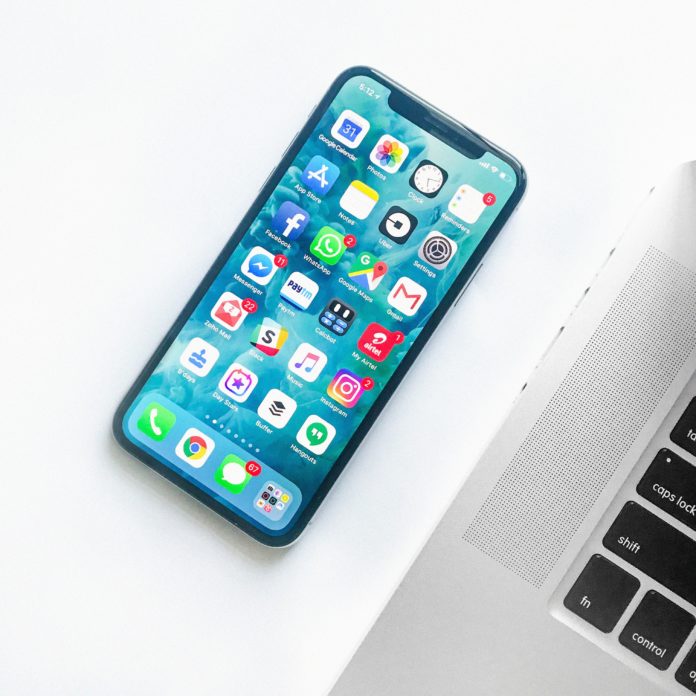 An iPhone is next to a computer keyboard. Multiple apps can be seen on the phone which has a blue background.