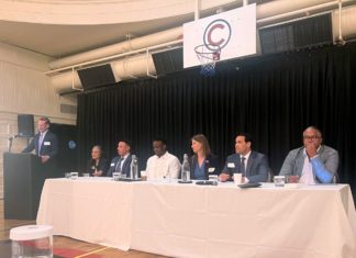 Coalition of Denver’s Five Largest Law Firms Marks Two Years of Efforts to Create a More Inclusive Bar A group of six people, men and women of diverse ages and races, in professional clothes sit in front of a table in a gym. On the far left, a man in a suit in front of a podium speaks.