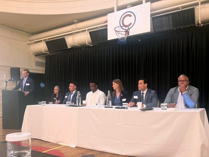 A group of six people, men and women of diverse ages and races, in professional clothes sit in front of a table in a gym. On the far left, a man in a suit in front of a podium speaks.