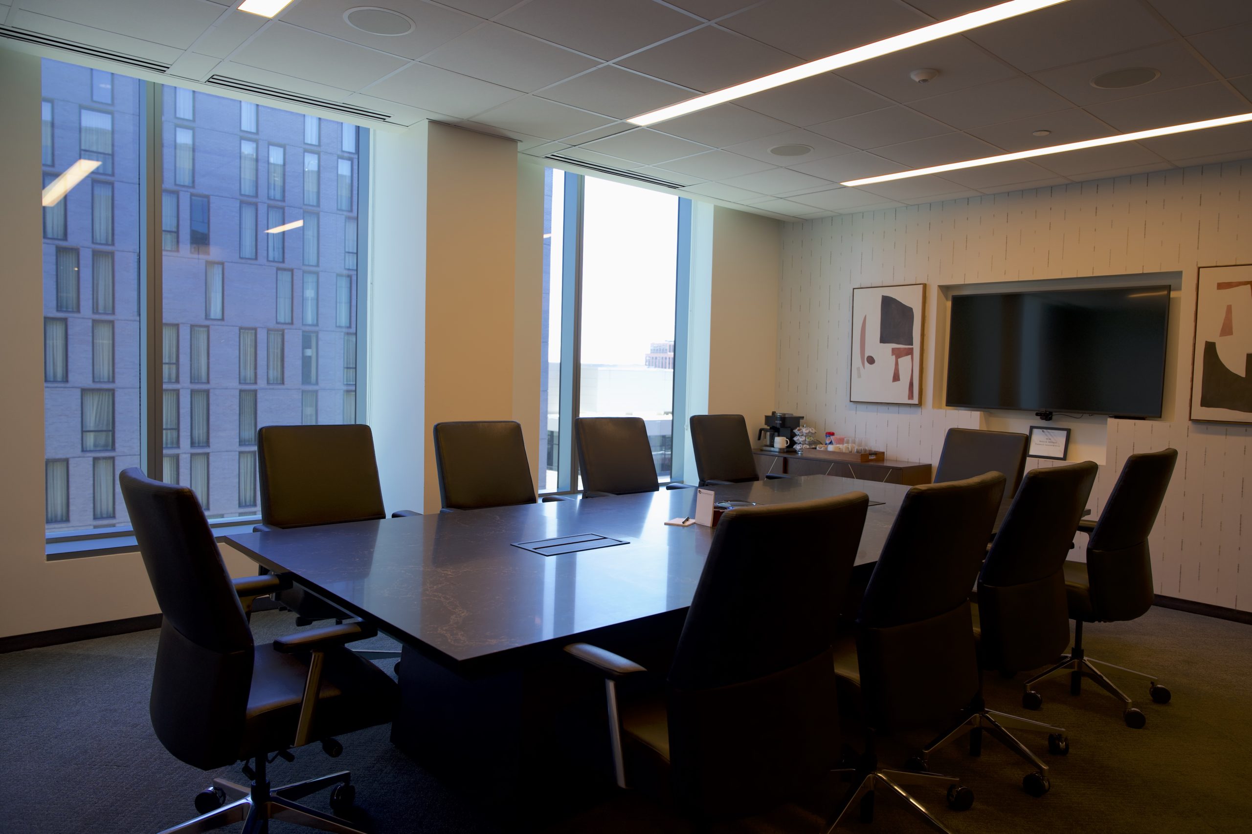 A modern office conference room. A large table with ten desk chairs around it. Large windows behind show office buildings. A TV and coffee station are on one wall. 