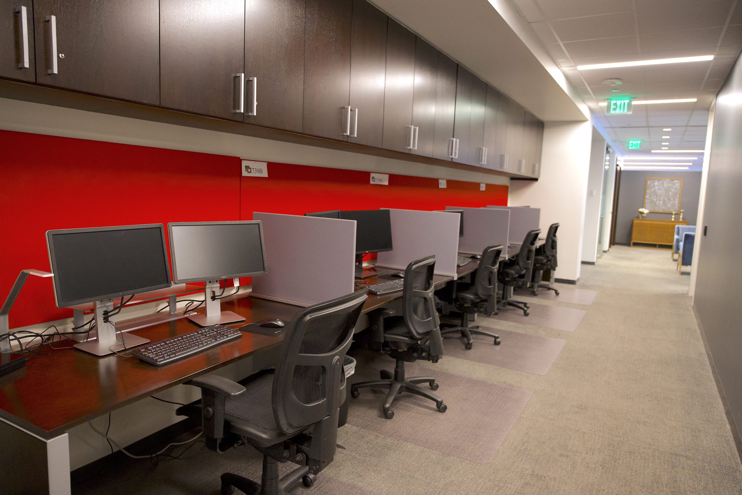 An office hallway with four desks and office chairs against one side with a red wall. Each desk is clean and has a keyboard and two monitors. 