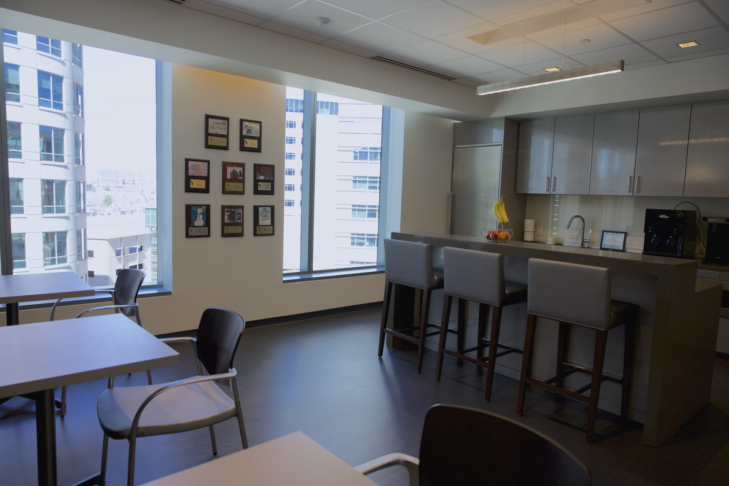 A modern office kitchen. Three small tables line one wall, a kitchen and counter with stools are on the other. Large windows show the downtown skyline outside. On a wall are eight small frames with children's drawings and plaques. 