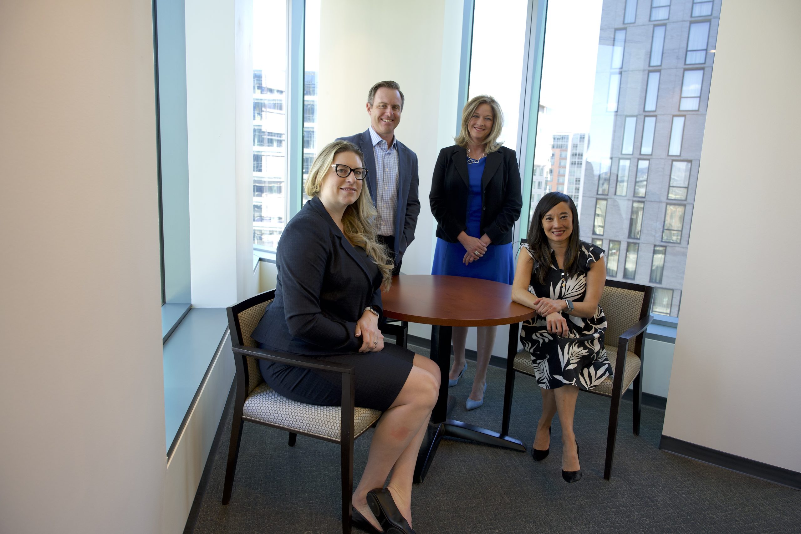 Four people dressed in professional clothes in an office building pose for a picture. On the left sitting at a small table is a blonde woman with black glasses in a suit. On the right of the table is an Asian woman with black hair in a patterned dress. Behind them on the left is a man with brown hair in a gray suit. On the right is a blonde woman in a blue dress and a black blazer. 