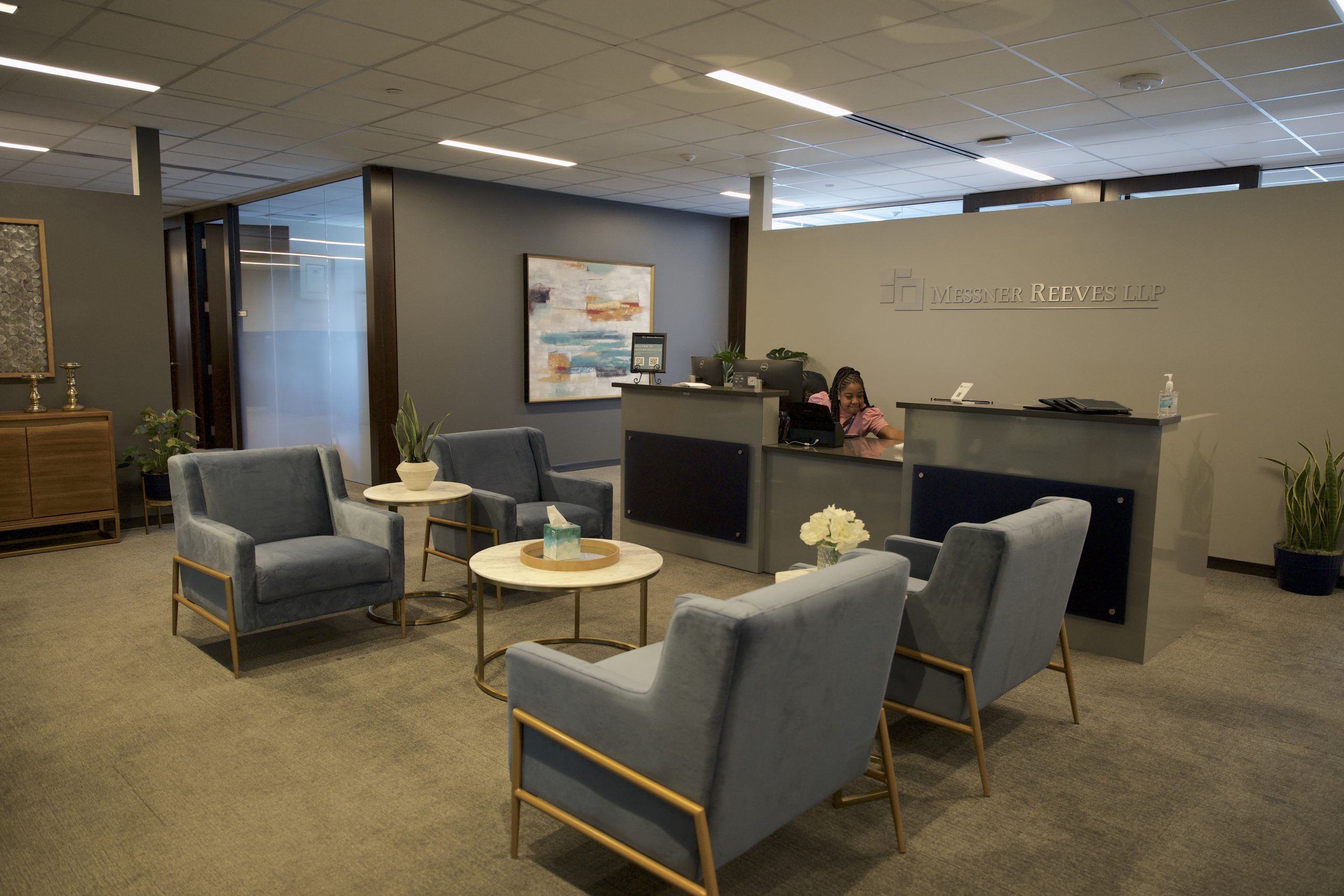 A modern office building lobby. Four blue chairs around white marble tables. Behind them is a reception desk with a Black woman in a pink shirt at the desk. Behind the desk is a chrome logo that reads Messner Reeves. 