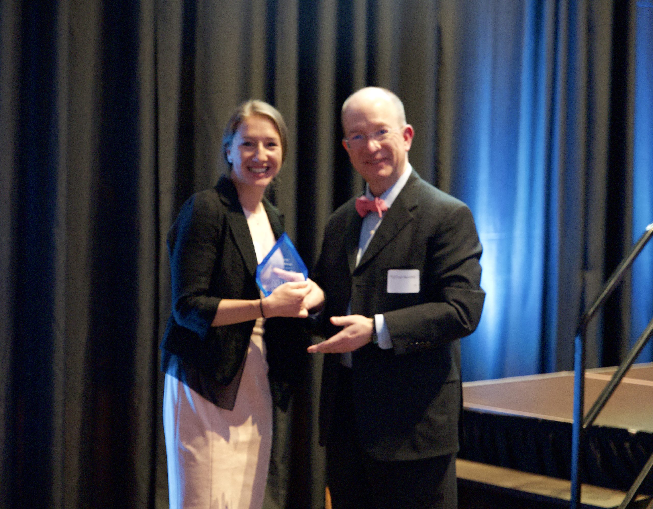 A blurry photo of two people standing close and smiling at the camera holding a crystal award. On the left is a younger woman with blonde hair in a black blazer and light pink dress. On the right, a middle aged bald man in a suit with a red bowtie smiles slightly. 