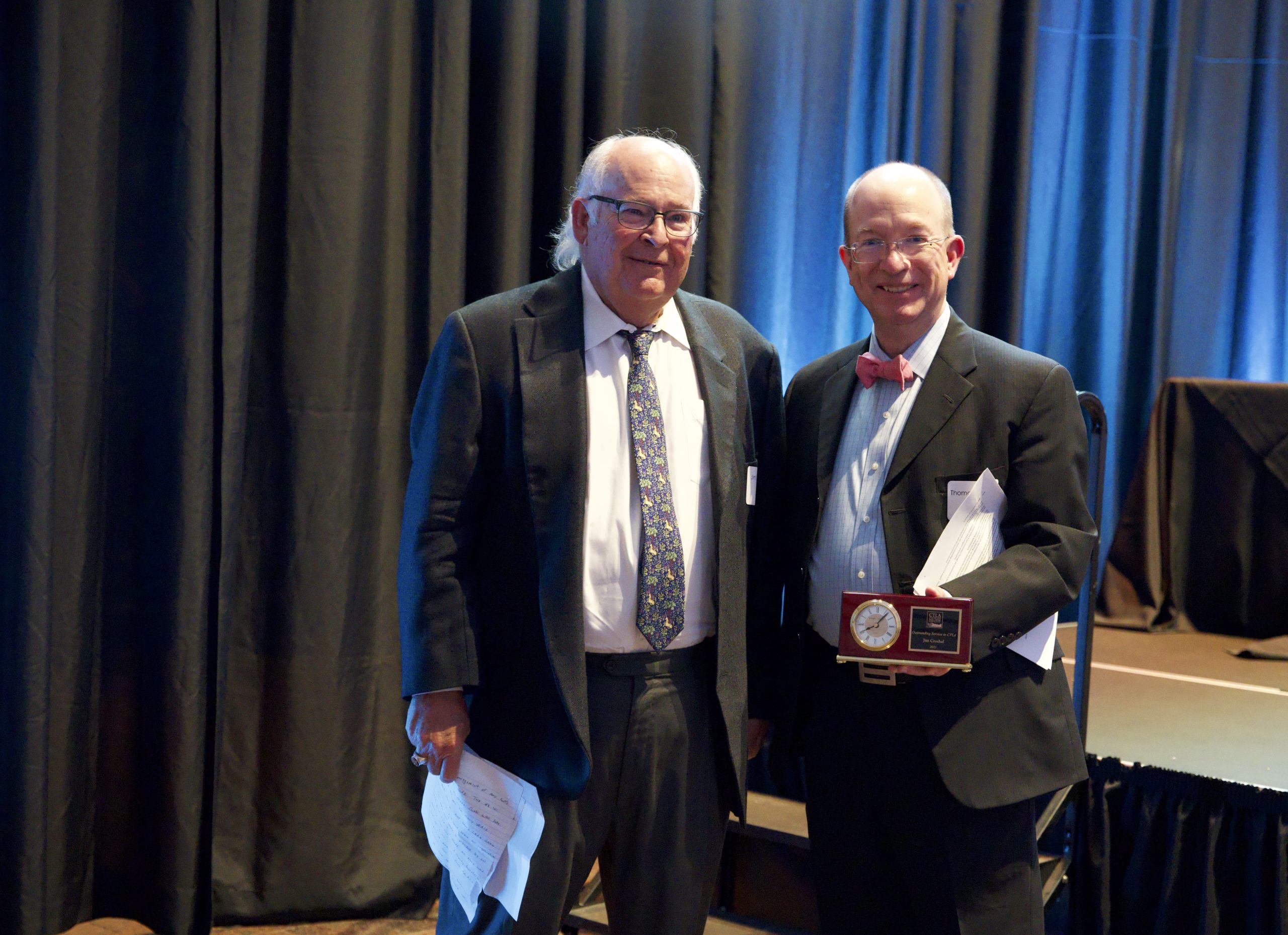 Two men pose for a picture together. On the right, an older man with long white hair wearing a suit holding papers. On the right, a middle aged bald man in a suit with a red bowtie smiles and holds a plaque with a clock embedded in it. 