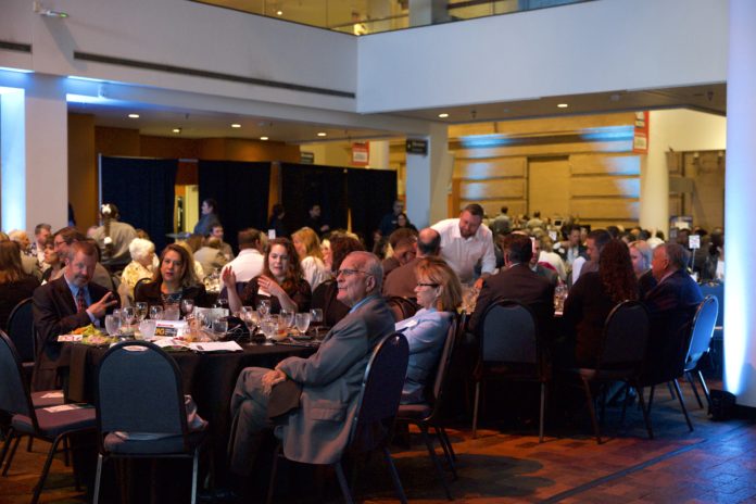 A wide shot of a formal banquet in a large atrium. People in formal clothes sit around round tables, some speaking to each other and others looking toward the left.