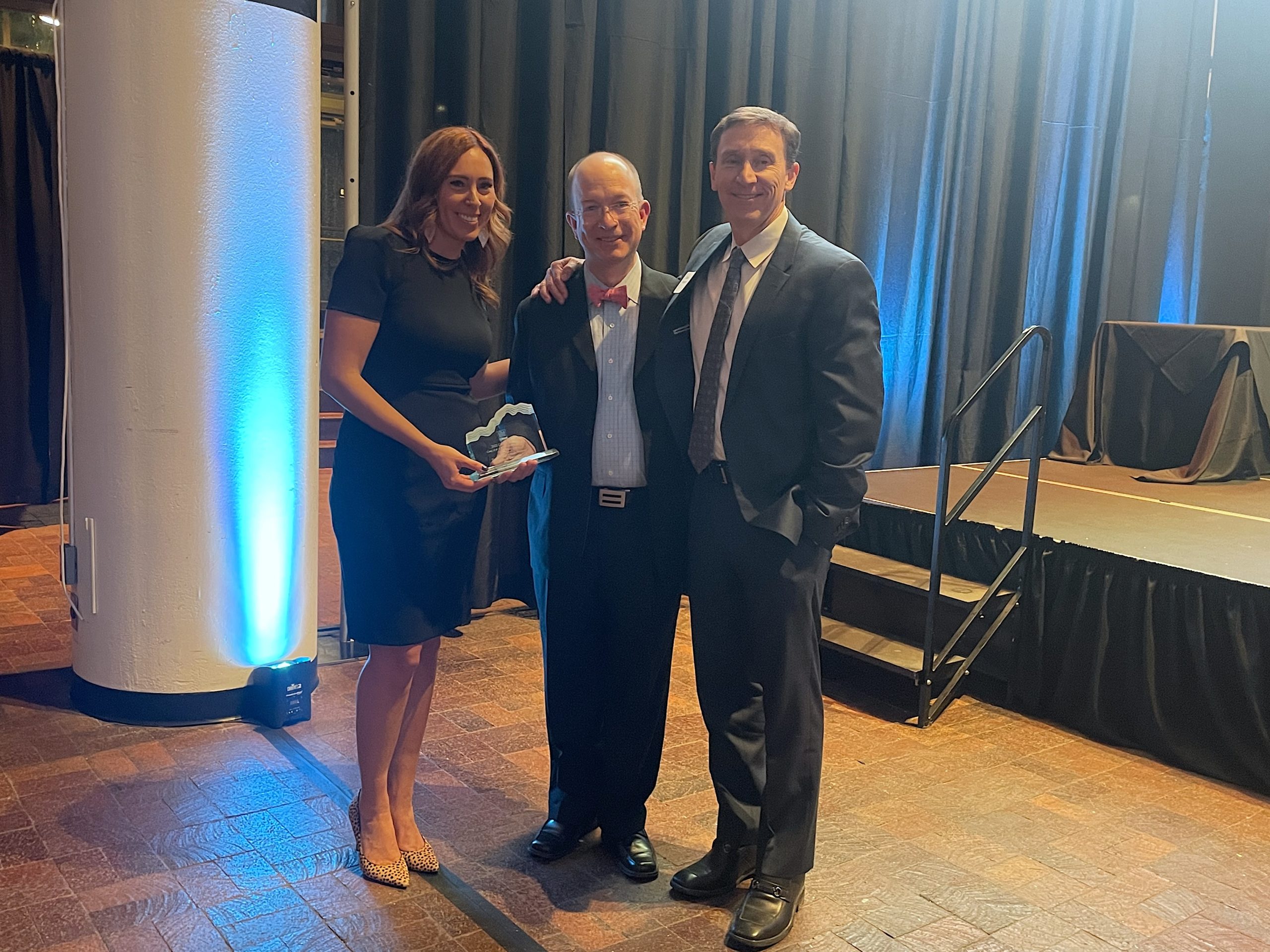 Three people smile for the camera while two of them hold a crystal award. On the left is a woman in a black dress with red hair. In the middle is a middle aged bald man in a suit with a red bowtie. On the right is a middle aged man with brown hair in a dark suit. 