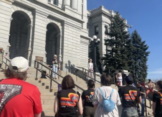 Colorado Public Defenders Join May Day Rally A group of people in colorful t-shirts watch a woman with short blonde hair in jeans speak on the steps of the Colorado capitol.